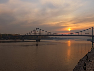 Pedestrian Bridge over Dnipro River during sunrise. The sun is reflected in the water. Men are fishing on the embankment. Autumn landscape. Kyiv, Ukraine