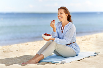 Fototapeta na wymiar people and leisure concept - happy smiling woman eating strawberries on summer beach