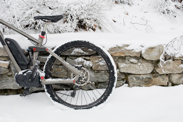 detail of electric bicycle, e-bike, ebike, resting on a wall of rocks, snowy road, mountain forest during snow storm, blizzard, sports, end of winter, wheel, cold, alps, Piedmont, Italy