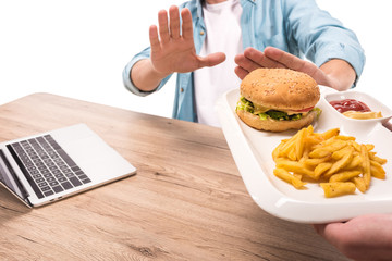 cropped image of man rejecting unhealthy burger and french fries at table isolated on white