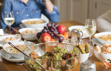 This is a table full of food, including various salads, pickles, soup, bread, fruit, glasses of white wine and a can of drink. People eat at the table.