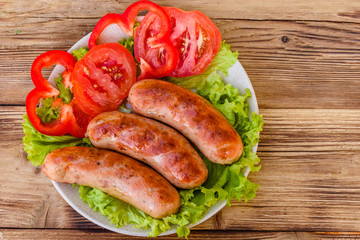 Ceramic plate with grilled sausages, sliced tomatoes and lettuce leaves on wooden table. Top view