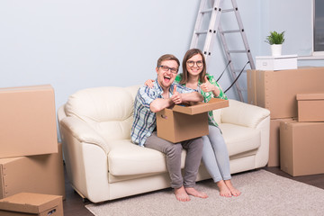 Real estate, relocation and moving concept - happy couple in glasses holding a card board and showing thumbs up