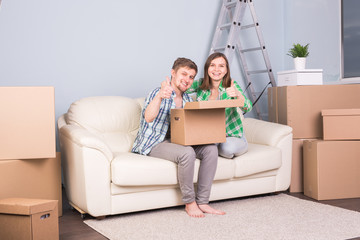 Real estate, relocation and moving concept - young couple holding a cardboard sitting on sofa and showings thumbs up