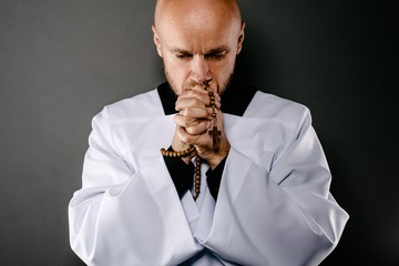Priest in white surplice and black shirt praying with wooden rosary
