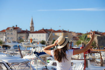 Young travelling woman in hat taking selfie photos over summer sunny mediterranean city with smartphone camera. Brunette girl making photography on summer vacation