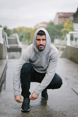 Sporty man getting ready for winter running and outdoor fitness workout under the rain. Sportsman lacing sport shoes on urban stairs.
