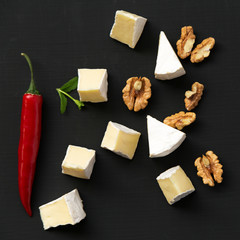 Slices of cheese camembert or brie  with walnuts and chili pepper over black background, overhead view. Milk production. Top view. Flat lay, from above.