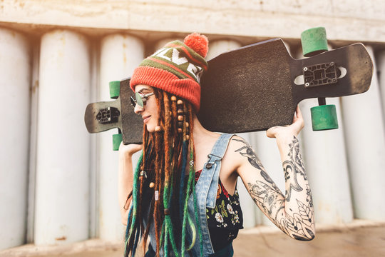 young girl in sun glasses and a cap stands with a longboard against the background of concrete structure