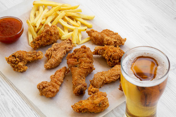 Fast food: fried chicken drumsticks, spicy wings, French fries and chicken fingers with sour-sweet sauce and cold beer over white wooden background, low angle view.