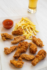 Tasty fastfood: fried chicken drumsticks, spicy wings, French fries and chicken fingers with sour-sweet sauce and cold beer over white wooden background, low angle view.