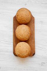 Fresh burger buns with sesame seeds on wooden board on white wooden table, overhead view. Fast food.