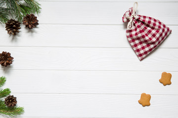 Christmas holidays background, copy space. Fir and pine tree branches with pinecones, christmas chequered red and white bags, ginger cookies on white wooden background, top view.