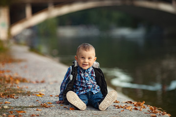 A little boy is sitting next to the yellow autumn leaves and lake during a walk in the park.