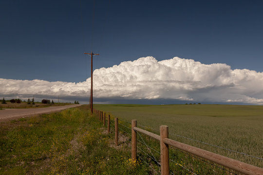 Huge Cumulonimbus cloud over farmland in Okotoks Alberta, view of a road lined with wooden post fences and hay and grass field.