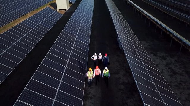 Aerial zoom shot of six electrical workers walking in between long rows of photovoltaic solar panels