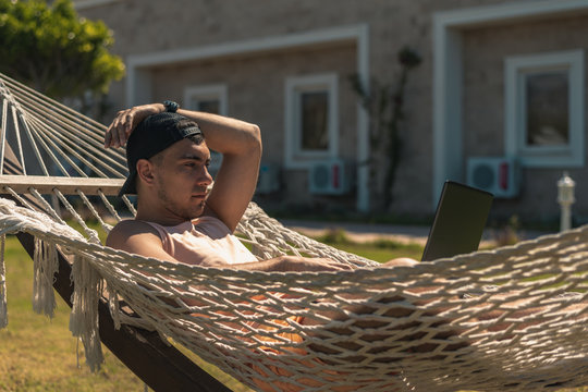 Young man in cap lying in hammock with laptop, remote work, freelance, work online