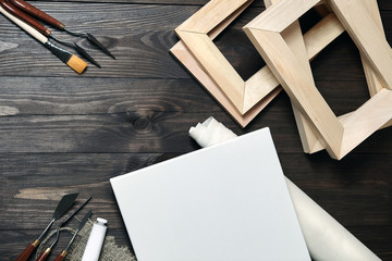 A white blank canvas, palette knives, synthetic paintbrush, a tube with oil or acrylic paint and stretchers lying on a brown wooden background. Top view