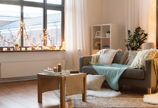 interior, christmas and interior concept - cushioned sofa, coffee table, garland string and candles on window sill in living room of cozy home
