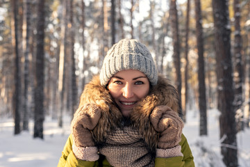 Fototapeta na wymiar Horizontal winter portrait of a young woman on a coniferous forest background on a Sunny day. Girl in a jacket with fur, knitted hat and mittens smiling at the camera