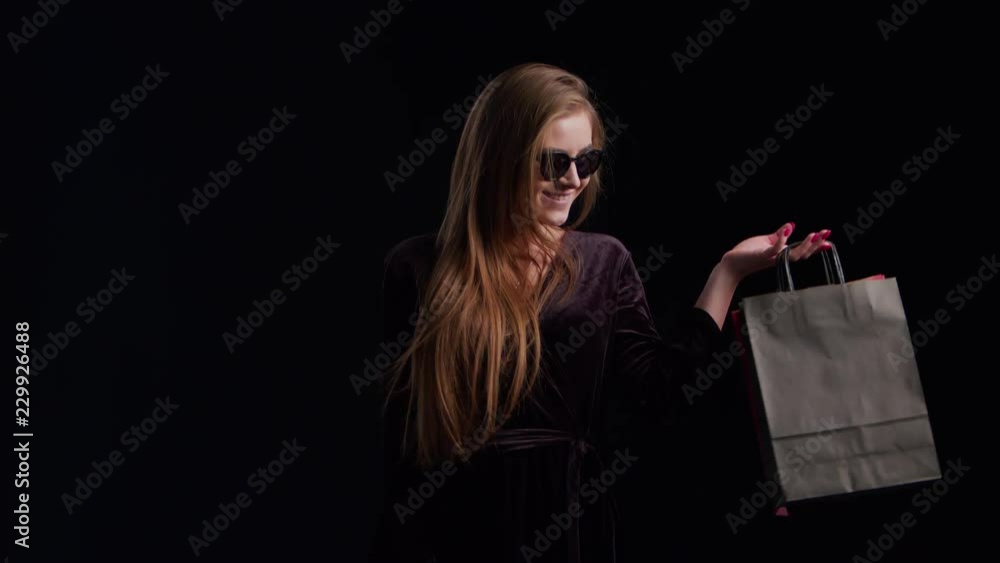 Wall mural Woman in cool sunglasses and black dress, holding black shopping bag isolated on dark background in black friday holiday or christmas