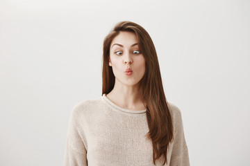 Obraz premium We all kids inside. Portrait of funny emotive adult female folding lips like fish and squinting to look at nose, being childish and carefree, having fun or fooling around, making faces over gray wall