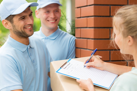 Smiling friendly couriers in blue uniforms and woman signing receipt of package delivery
