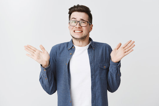 Sorry I Messed Up. Guilty Young And Handsome Male In Glasses And Blue Shirt Raising Hands Near Shoulders, Shrugging Making Awkard Smile As Apologizing For Making Mistake Over White Background