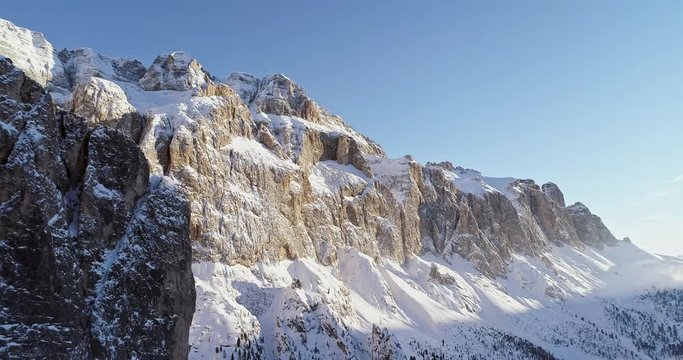 Backward aerial along snowy alpine steep rocky cliff valley.Sunny day,clear sky.Winter Dolomites Italian Alps mountains outdoor nature establisher.4k drone flight