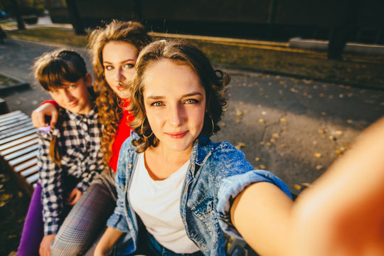 Happy three teen different ages girls friends taking selfie in park with mobile phone. Group of students looks at camera while relaxing on the bench in university campus.