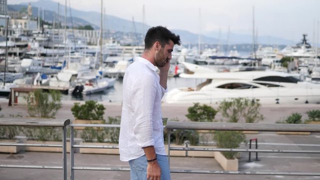 Athletic man in white shirt at the seaside in Montecarlo, Monaco's harbor, using cell phone to call someone with the sea behind him