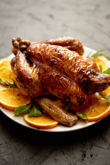 Roasted whole chicken or turkey served in white ceramic plate with oranges and lamb's lettuce on dark stone background. View from above.