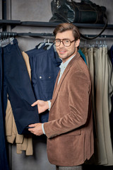 handsome young man in eyeglasses choosing clothes and smiling at camera in shop