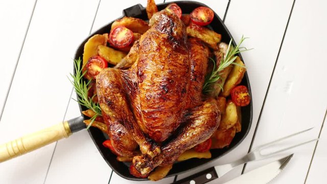 Roasted whole chicken in cast iron black pan. With potatoes, tomatos and rosemary herb. Placed in white wooden table. View from above. Shot with copy space.