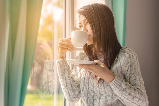 Asian woman in bedroom drinking coffee after wake up near window, sunny morning. Lifestyle Concept.