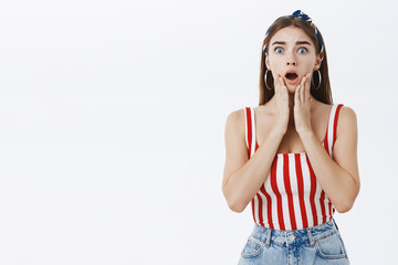 Waist-up shot of shocked girlfriend standing in stupor over grey background dropping jaw gasping from worry and shock holding palms on chin being scared of bad news looking concerned at camera