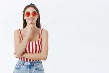 Girl thinking it perfect time to go travel. Portrait of creative happy and thoughtful good-looking female in sunglasses and striped top touching lip gazing up while making decision against grey wall