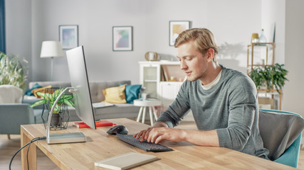 Portrait of the Focused Young Man Typing on a Personal Computer while Sitting at His Desk. In the Background Cozy Living Room.
