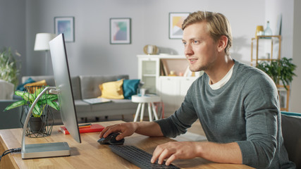 Portrait of the Focused Young Man Working on a Personal Computer while Sitting at His Desk. In the Background Cozy Living Room.