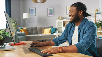 Portrait of the Smiling Stylish African American Working on a Personal Computer while Sitting at His Desk at Home.