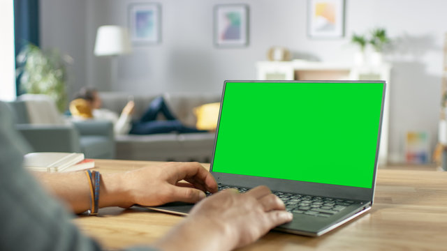 Over the Shoulder Shot of Mans Hands Typing on a Laptop with Green Mock-up Screen. In the Background Cozy Living Room with Woman Relaxing on a Sofa.