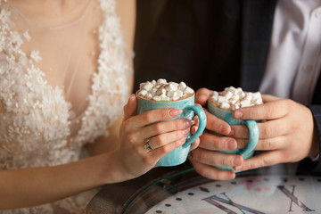 Wedding in cold weather. Bride and groom holding cups of coffee with marshmallow in cafe
