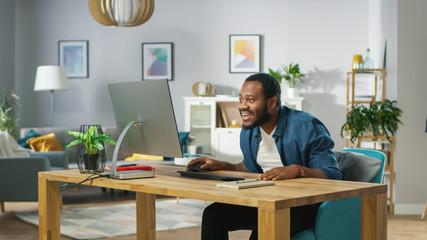 Shocked and Surprised Young Black Man Uses Personal Computer at Home. In the Background Cozy Living Room.