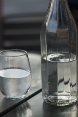 Faceted bottle and a glass are on a table. Both are half full with clean drinking water. Different shades of elegant gray.