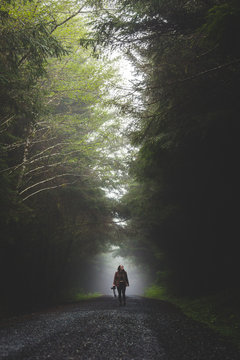 A young woman walks towards the camera down a foggy road in the forest