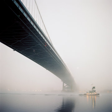 A river tugboat glides under a suspension bridge on a foggy evening.