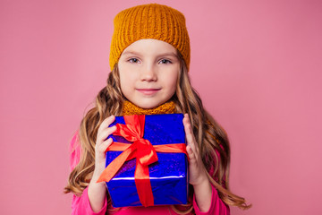 Happy little smiling blonde curly hairstyle girl in knitted orange hat with christmas gift box with bow on pink background studio. new Year present in hands of a female child making a wish copyspace