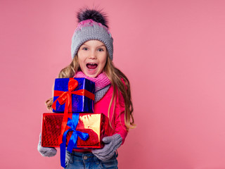Happy little blonde curly hairstyle girl in knitted pink gray hat ,scarf and mittens with christmas gift box pink background studio. new Year present in hands of a female child making a wish copyspace