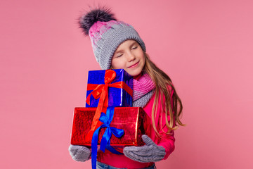 Happy little blonde curly hairstyle girl in knitted pink gray hat ,scarf and mittens with christmas gift box pink background studio. new Year present in hands of a female child making a wish copyspace