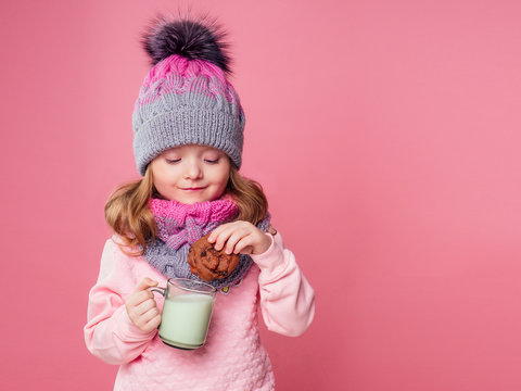 beautiful little girl drinks milk and eats cookies in a knitted hat and scarf on a pink background in the studio,christmas night